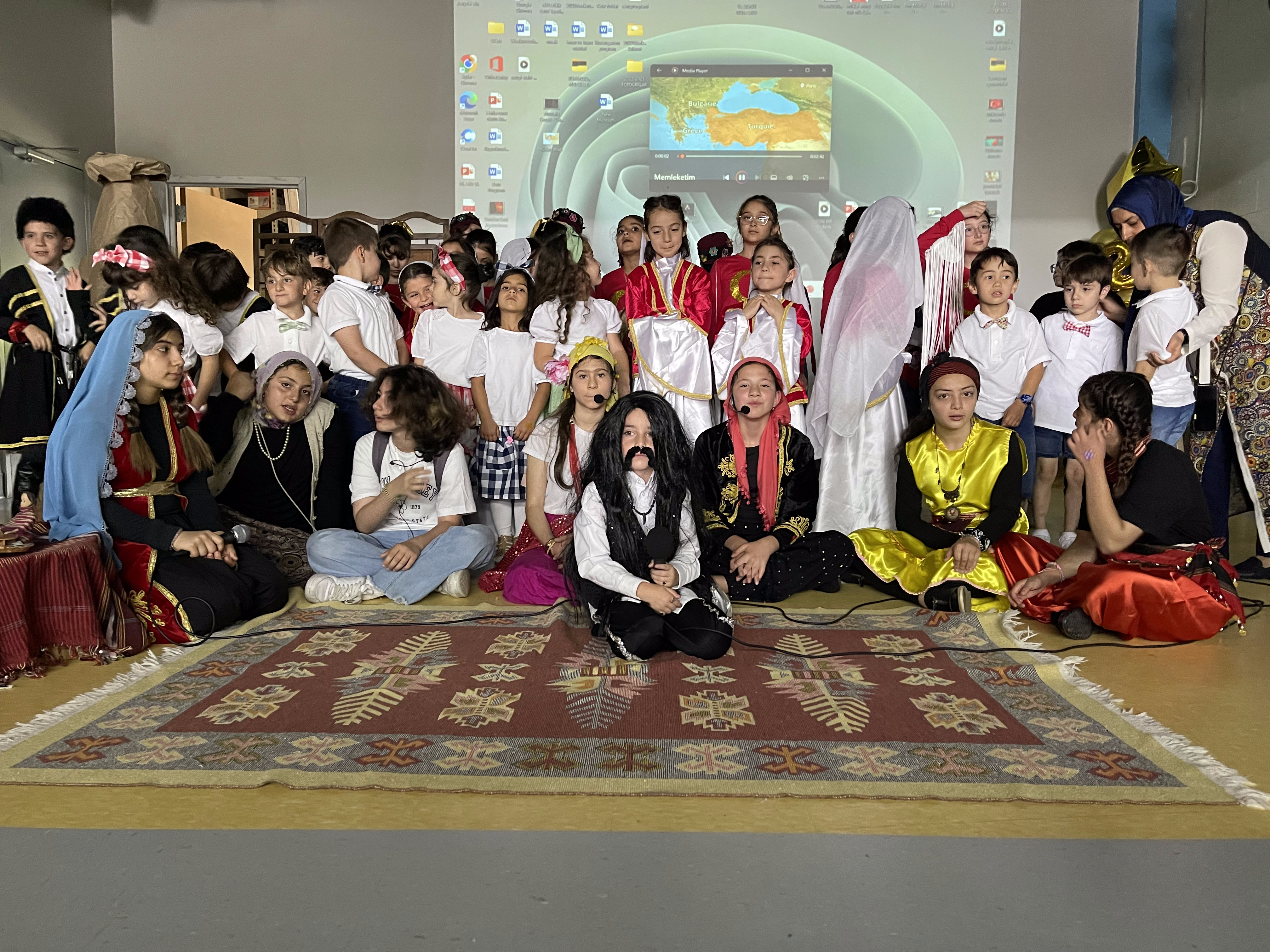 A large group of children in traditional costume seated together on a patterned carpet